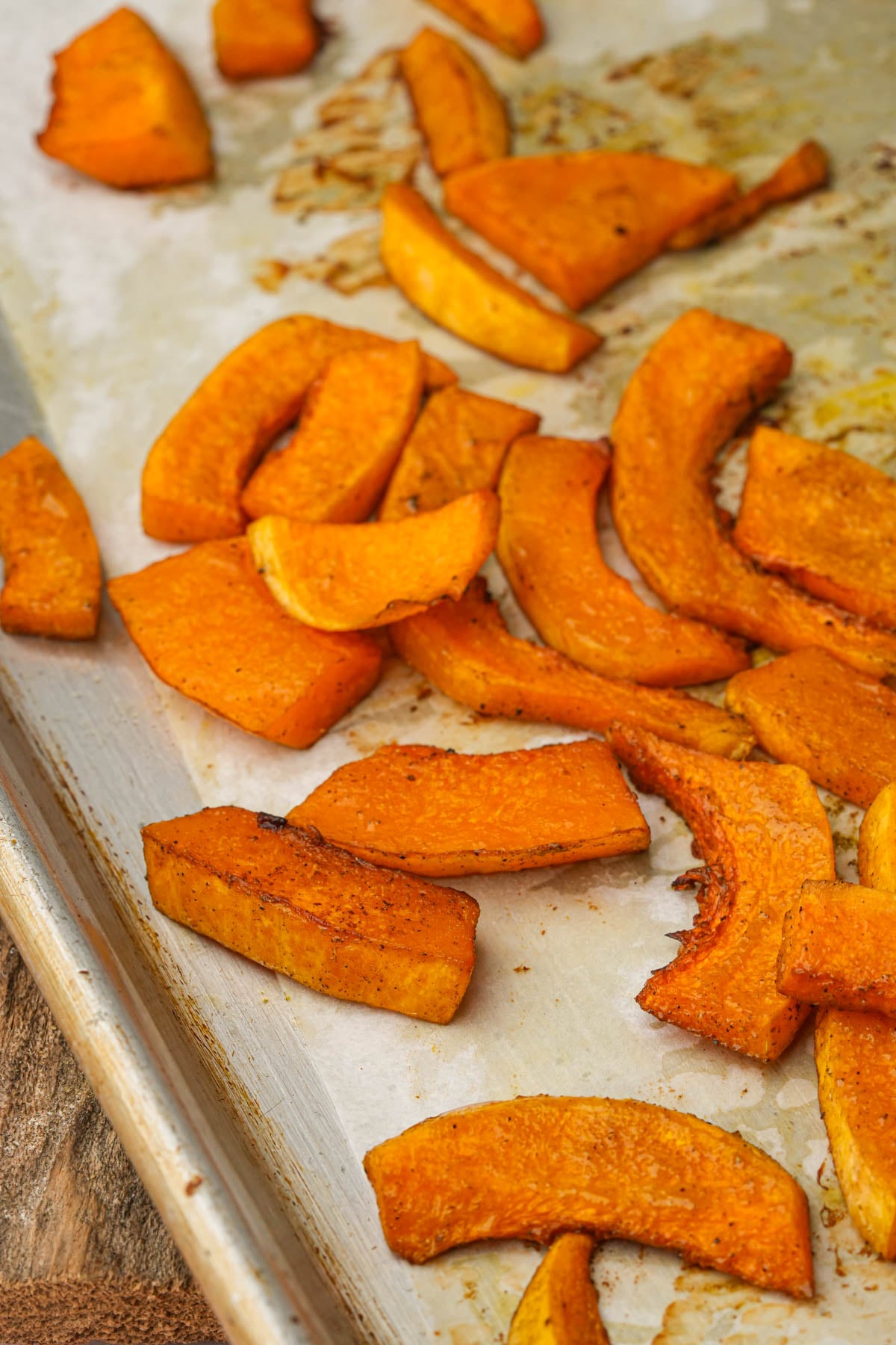 Slices of roasted butternut squash on a baking sheet, seasoned and slightly browned, with some pieces spread out and others grouped together.