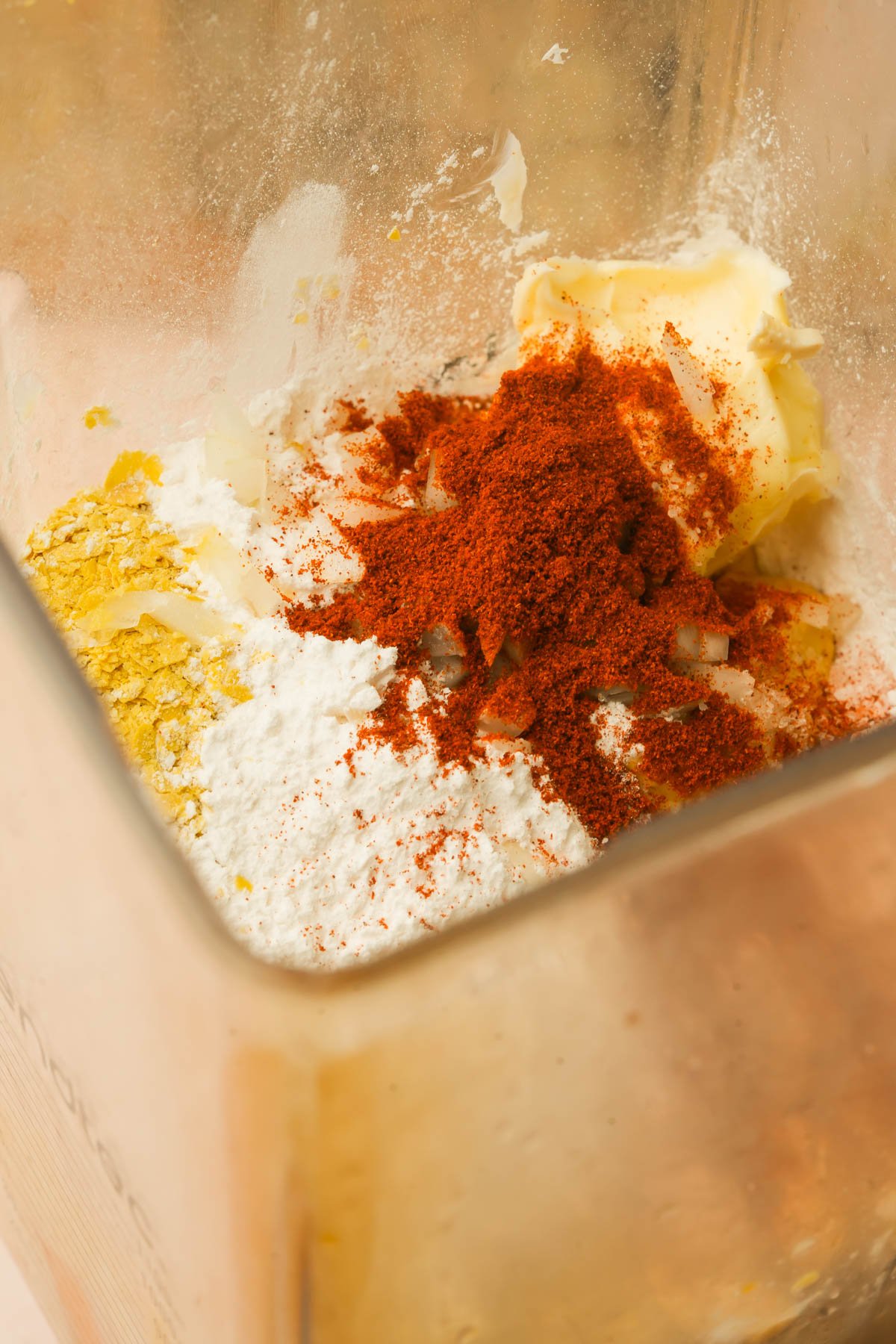 A close-up view of a blender container with vegan butter, paprika, nutritional yeast, and powdered ingredients ready to be blended together.