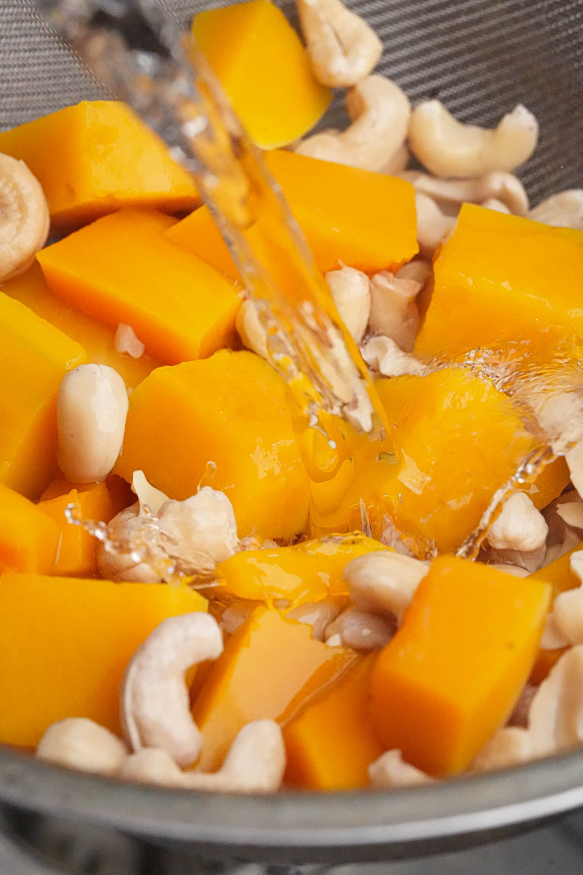 Chunks of bright orange butternut squash and cooked cashews in a metal bowl with a stream of water being poured over them.