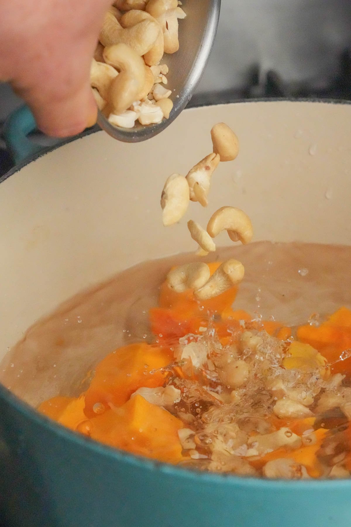 A hand pours cashew nuts from a small bowl into a pot of boiling water with orange butternut squash chunks.