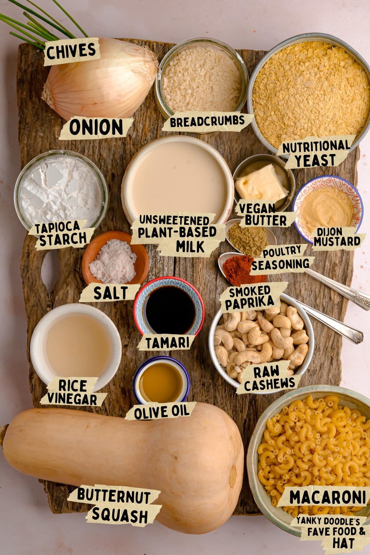 A wooden board with labeled ingredients for a vegan butternut squash macaroni and cheese recipe, including butternut squash, macaroni, onion, chives, raw cashews, plant-based milk, spices, oil, nutritional yeast, and more.