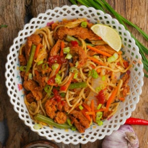 A bowl of stir-fried pancit canton noodles with sliced vegetables, strips of cooked soy curls, and garnished with chopped green onions, red sauce, and a lime wedge, served on a rustic wooden surface.