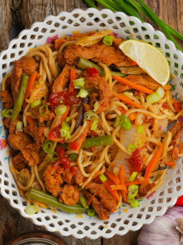 A bowl of stir-fried pancit canton noodles with sliced vegetables, strips of cooked soy curls, and garnished with chopped green onions, red sauce, and a lime wedge, served on a rustic wooden surface.