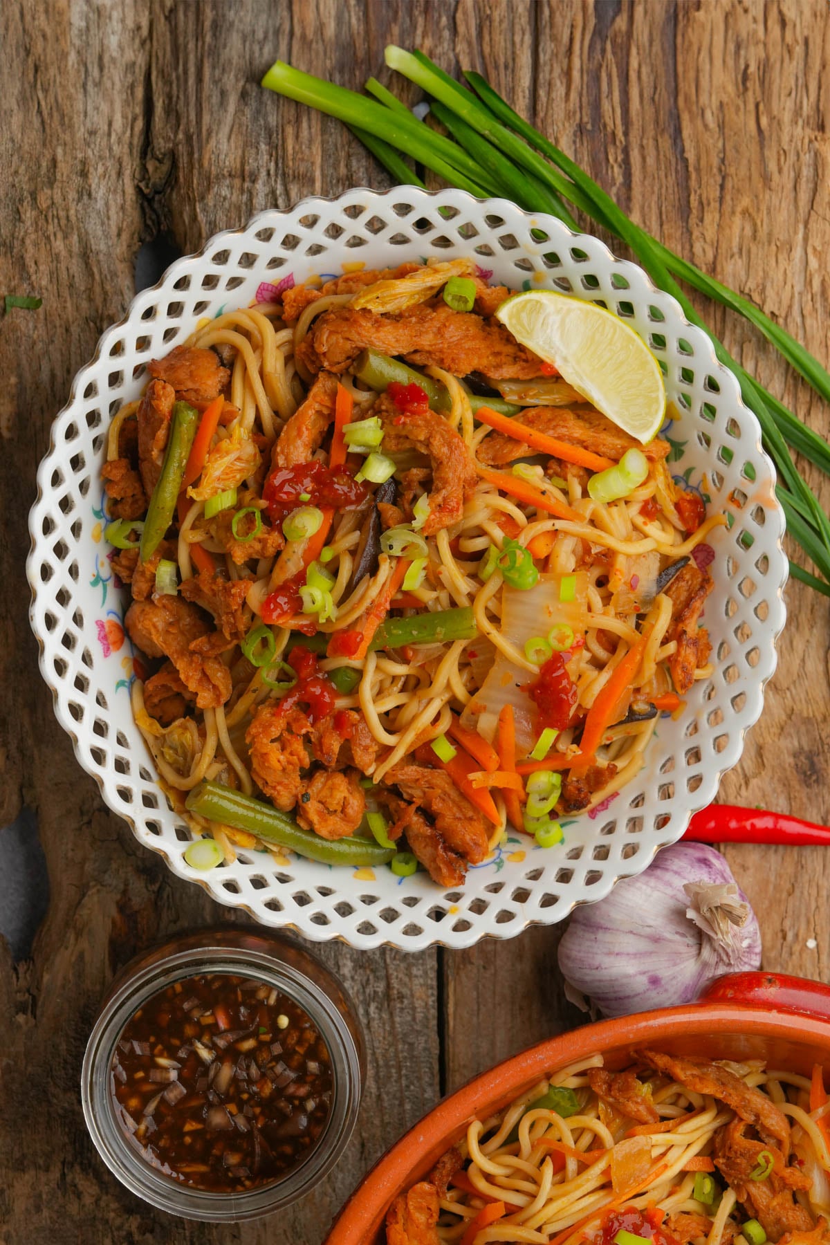 A bowl of stir-fried pancit canton with vegetables, shredded mock meat, scallions, a lime wedge, and chili sauce. Green onions, garlic, and a dipping sauce are placed nearby on a rustic wooden surface.