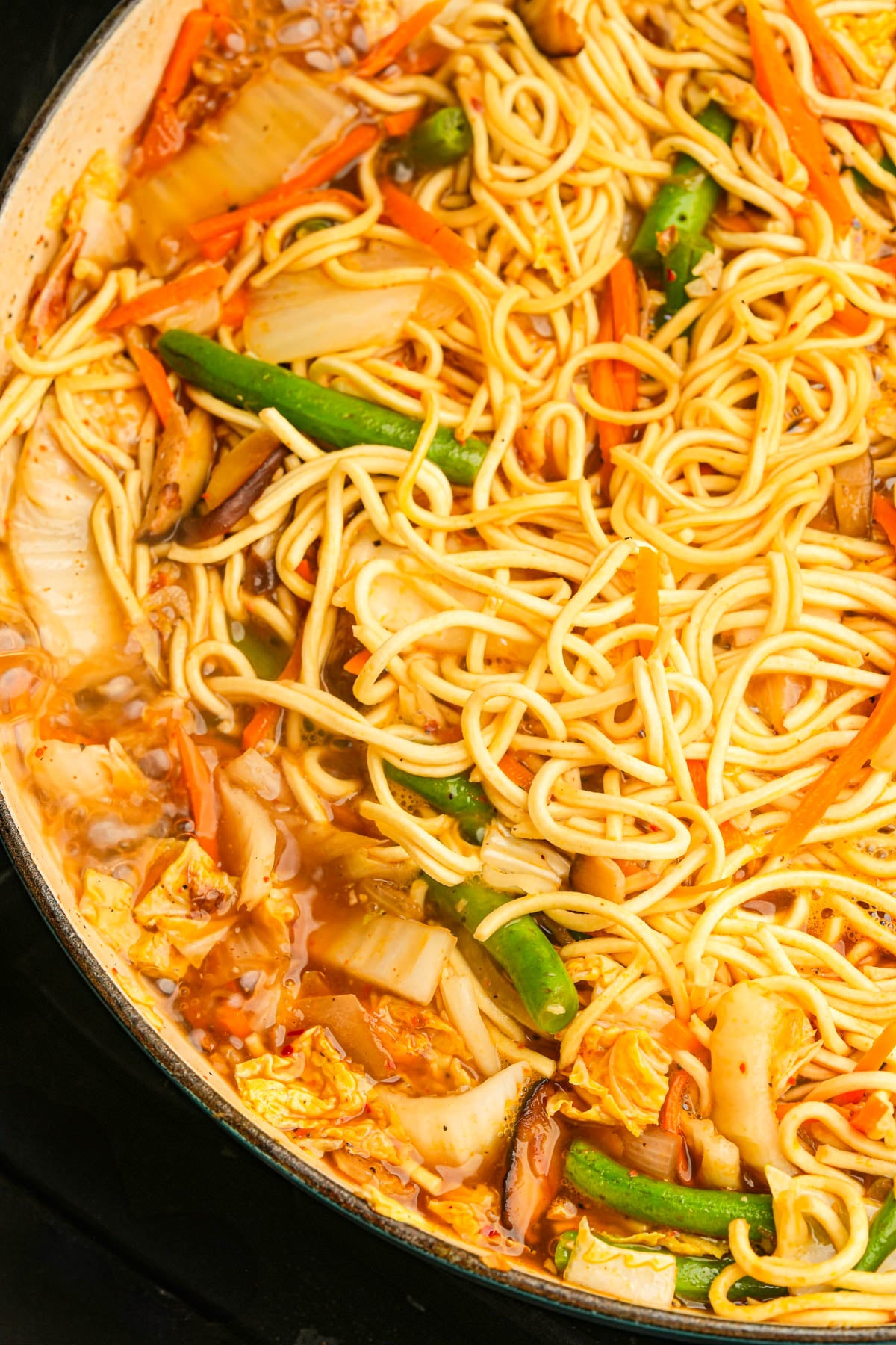 A close-up of a pot filled with cooking pancit canton with yellow noodles, cabbage, green beans, carrots, mushrooms, and a savory broth.