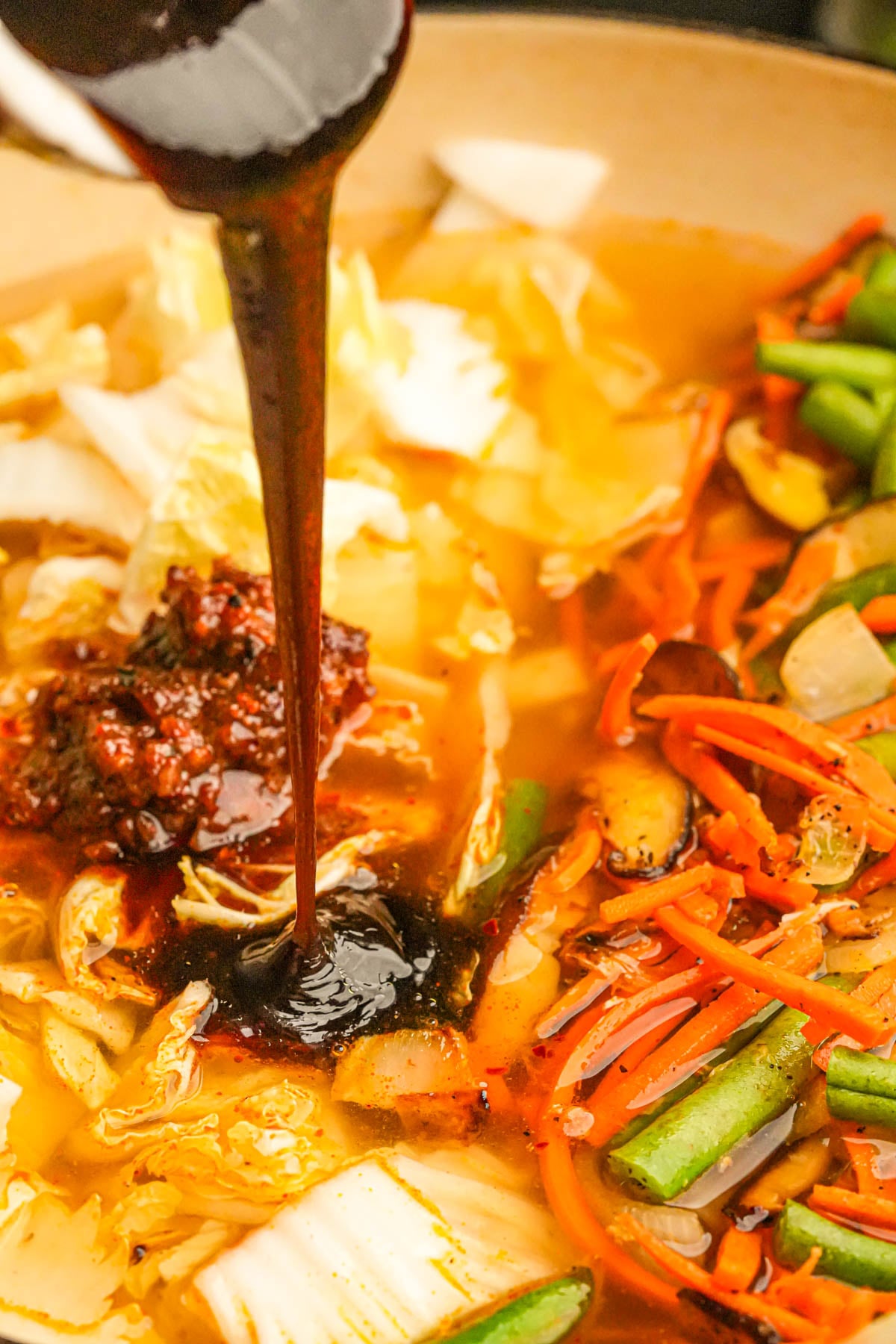 A close-up of a pot filled with assorted vegetables and broth, with a thick, dark sauce being poured in, creating a rich and flavorful soup or stew.