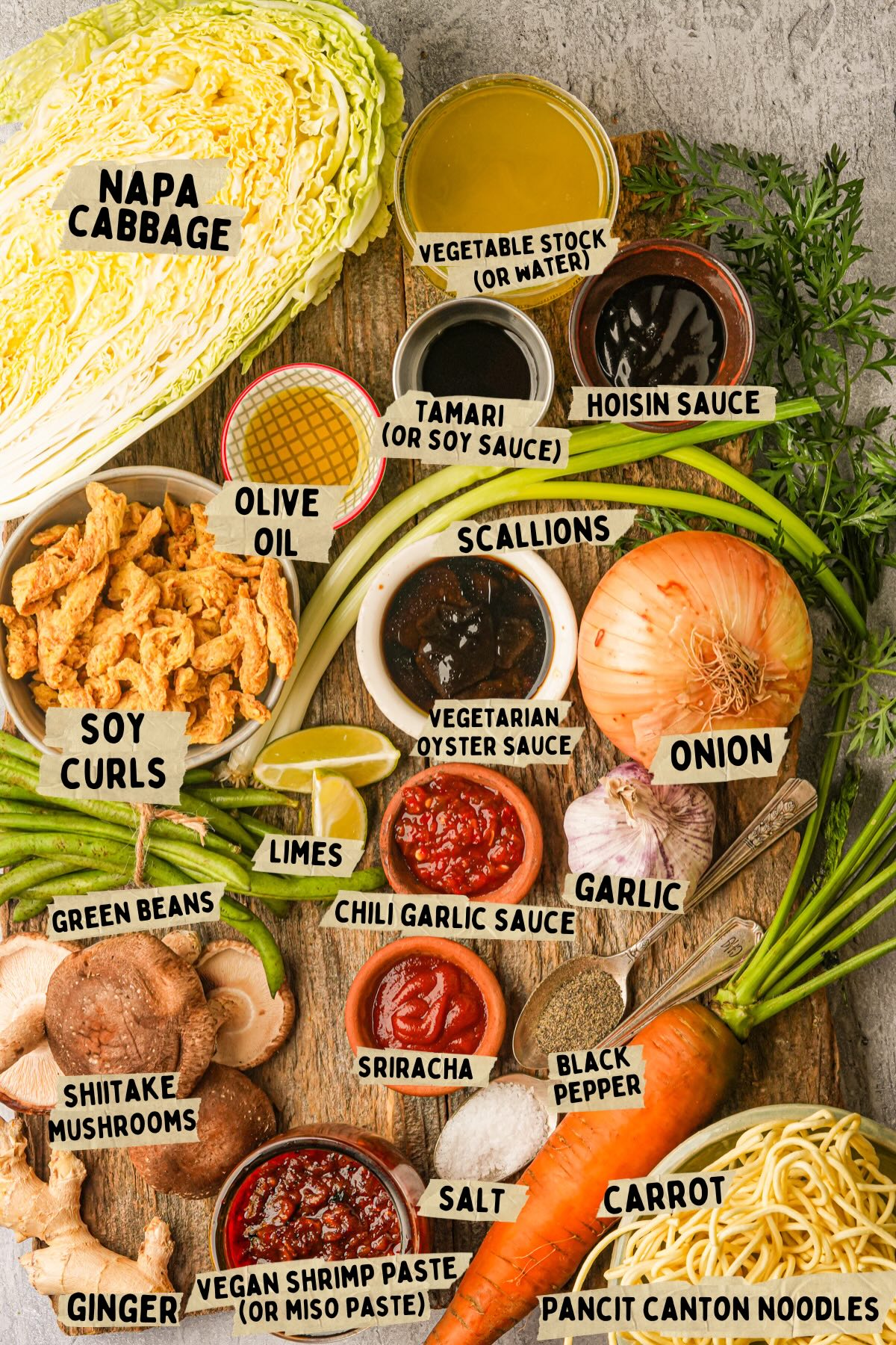 A variety of labeled vegan pancit canton ingredients, including napa cabbage, soy curls, shiitake mushrooms, carrot, onion, scallions, garlic, limes, sauces, ginger, green beans, and noodles, arranged on a wooden board.