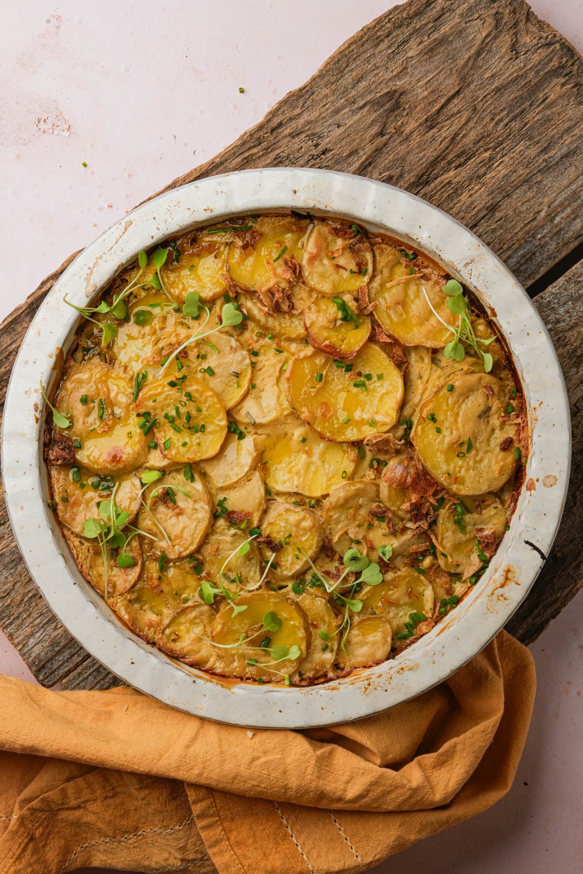 A round baking dish filled with golden, baked vegan scalloped potatoes, garnished with fresh herbs, sits on a rustic wooden board with a tan cloth nearby.