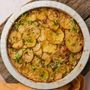 A round pan of vegan scalloped potatoes topped with thinly sliced golden potatoes, green herbs, and microgreens in a metal dish on a rustic wooden surface.