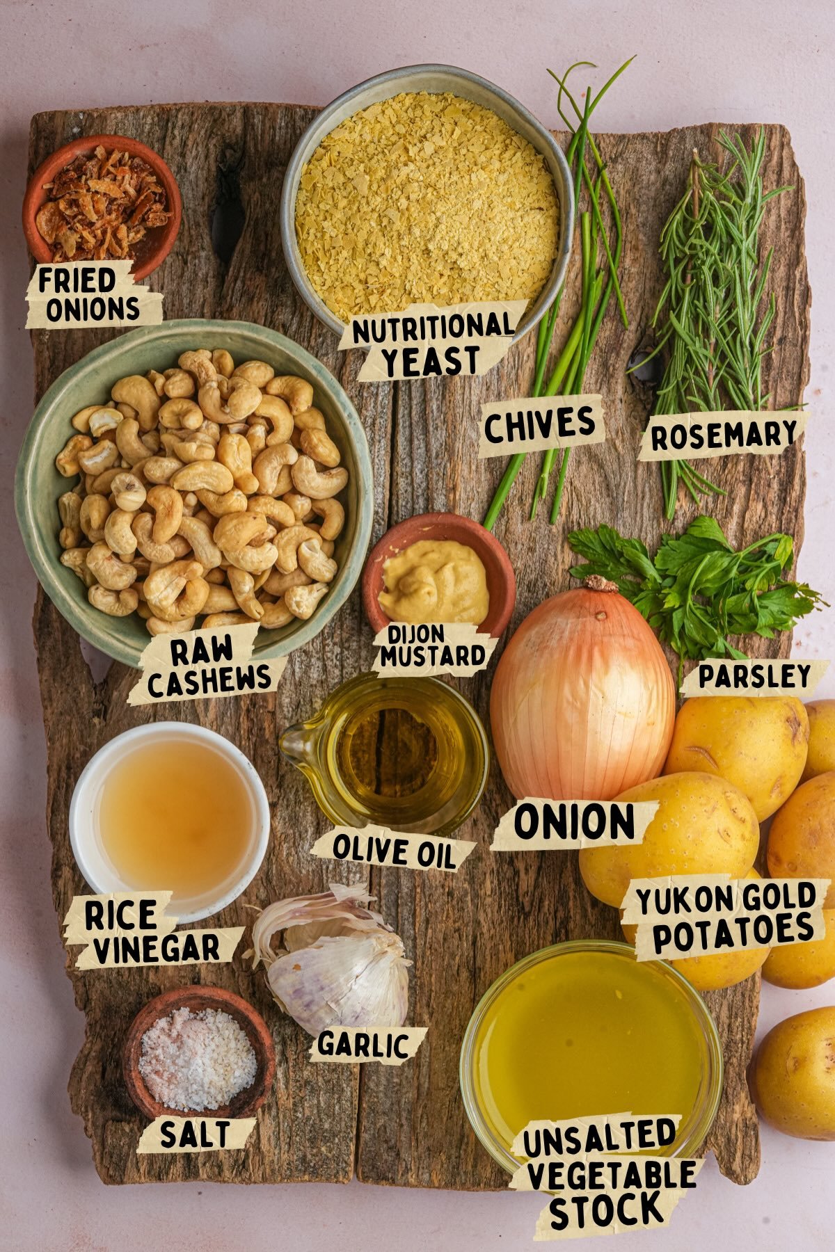 A rustic wooden board topped with labeled ingredients for a vegan scalloped potatoes recipe: fried onions, nutritional yeast, chives, rosemary, raw cashews, Dijon mustard, olive oil, parsley, an onion, Yukon Gold potatoes, rice vinegar, salt, garlic, and unsalted vegetable stock.