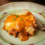 A slice of vegan shepherd’s pie topped with golden-brown gravy and chopped chives sits on a decorative plate with a fork beside it.