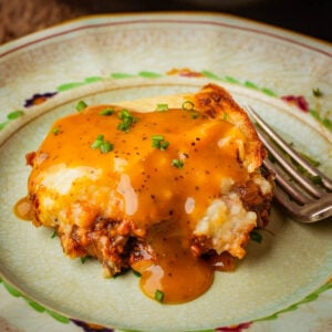 A slice of vegan shepherd's pie topped with golden-brown gravy and chopped chives sits on a decorative plate with a fork beside it.