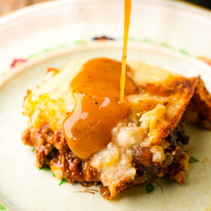 A close-up of a slice of vegan shepherd's pie with rich brown gravy being poured over the mashed potato topping, revealing layers of savory ground meat and vegetables on a plate.