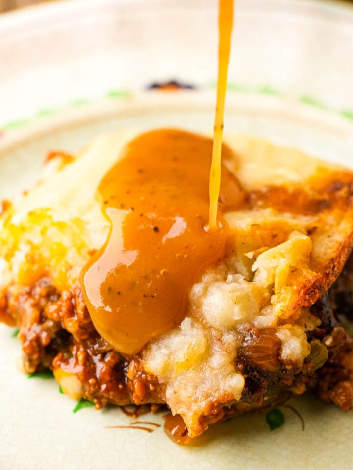 A close-up of a slice of vegan shepherd’s pie with rich brown gravy being poured over the mashed potato topping, revealing layers of savory ground meat and vegetables on a plate.