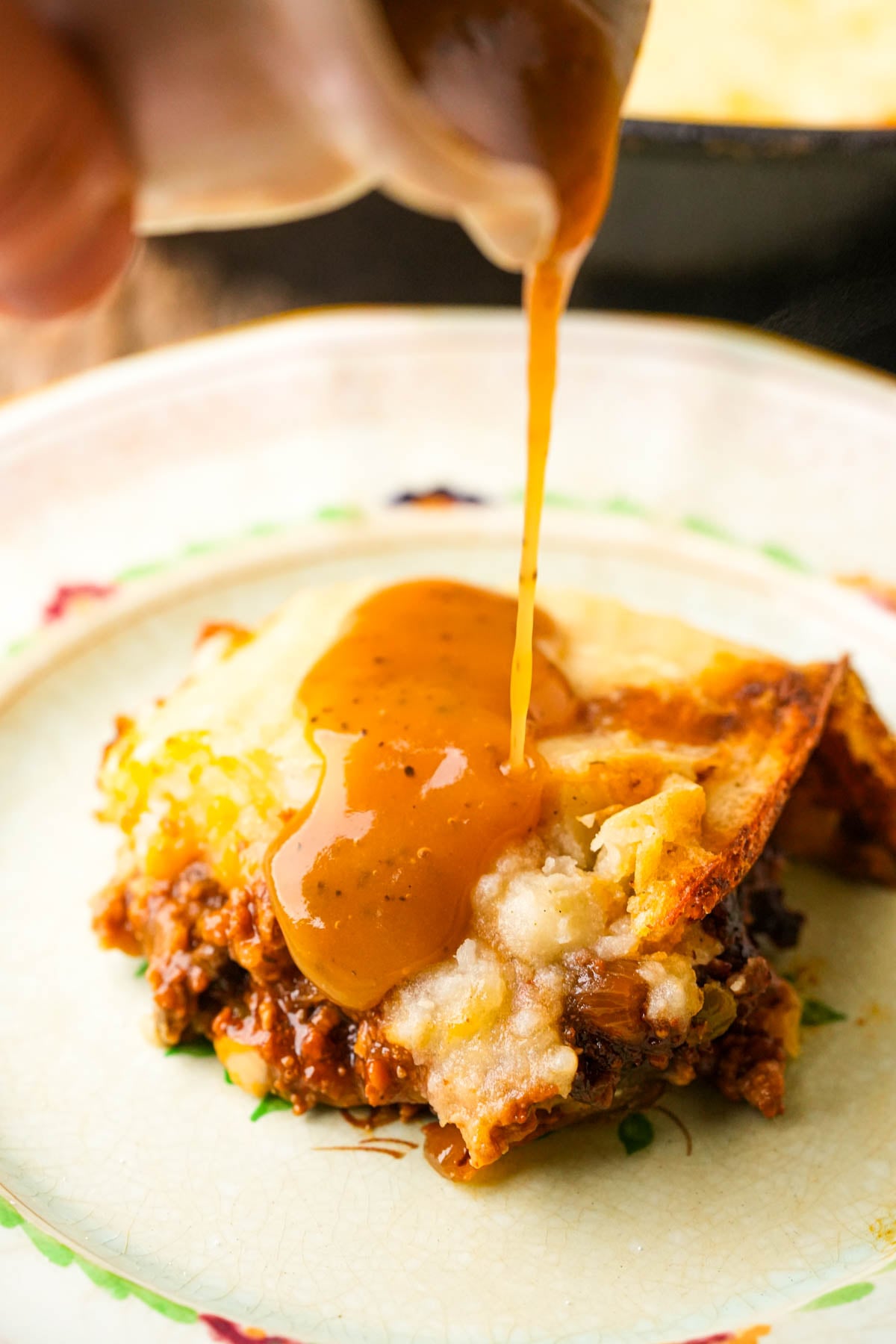Gravy being poured onto a serving of vegan shepherd's pie on a decorative plate, showing layers of mashed potatoes and seasoned meat filling.