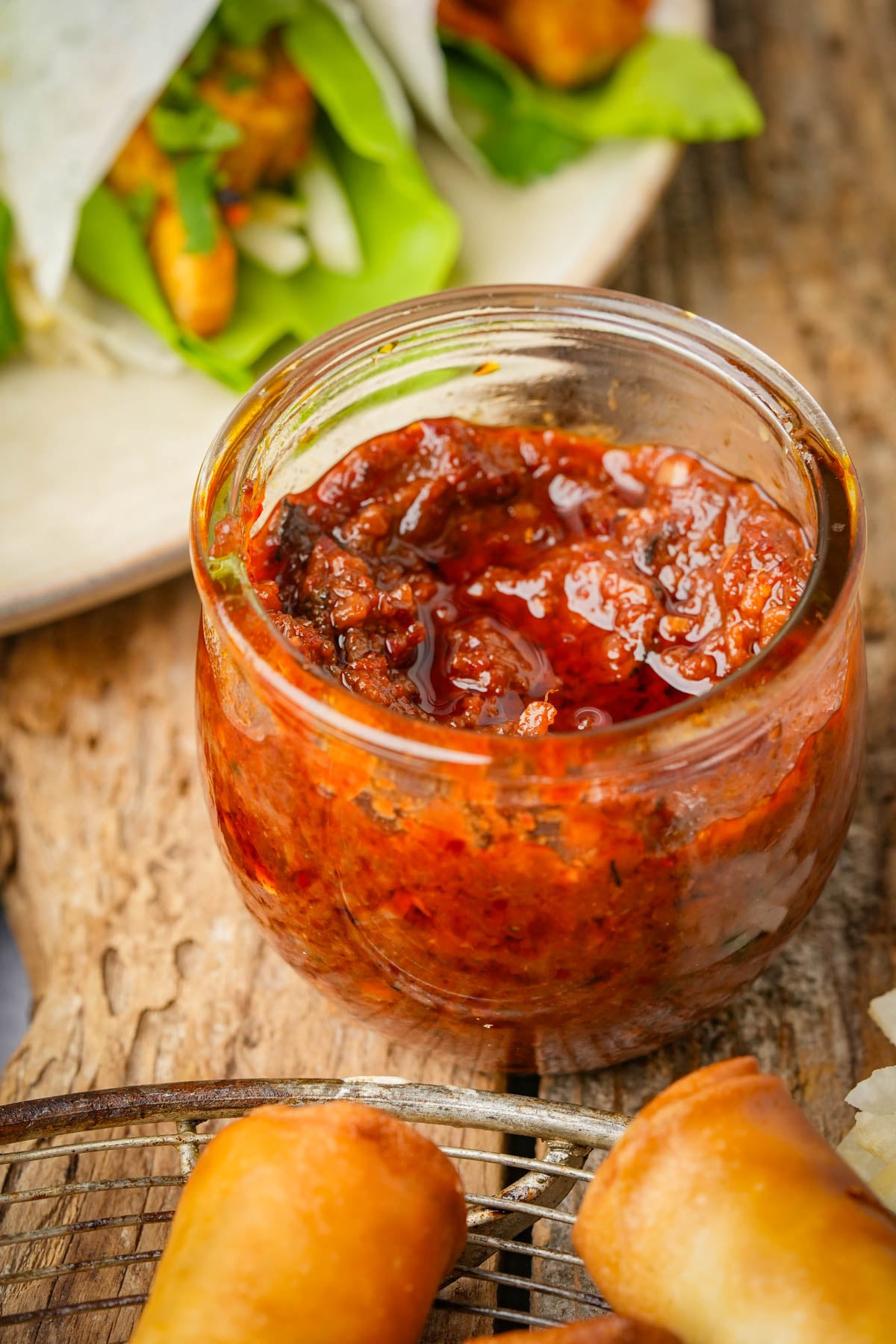 A glass jar filled with chunky vegan shrimp paste on a rustic wooden surface, surrounded by spring rolls and a plate of fresh greens and vegetables in the background.