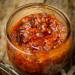 A close-up of a glass jar filled with chunky, red vegan shrimp paste placed on a rustic wooden table.