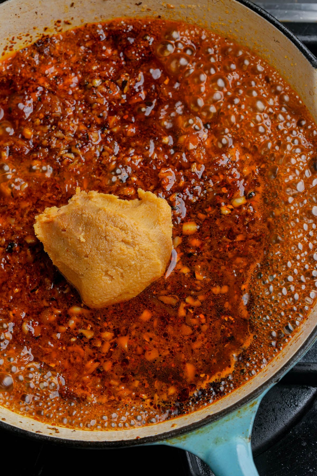 A close-up of a pan filled with bubbling red chili oil, minced garlic, and a large dollop of miso paste in the center, cooking on a stovetop.
