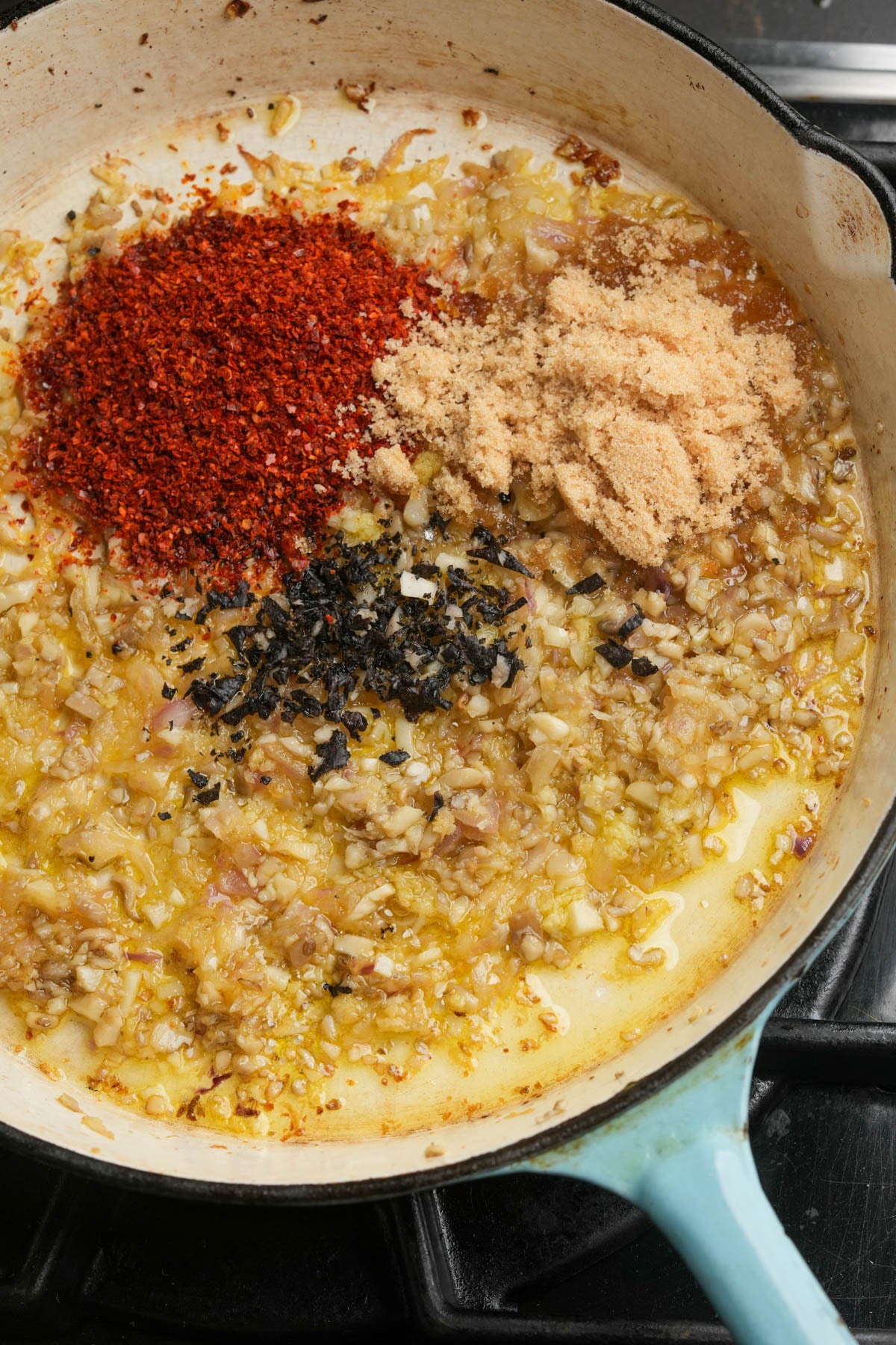 A close-up of a pan on a stove with chopped onions and garlic sautéing in oil, topped with piles of red chili flakes, brown sugar, and nori bits, ready to be mixed together.