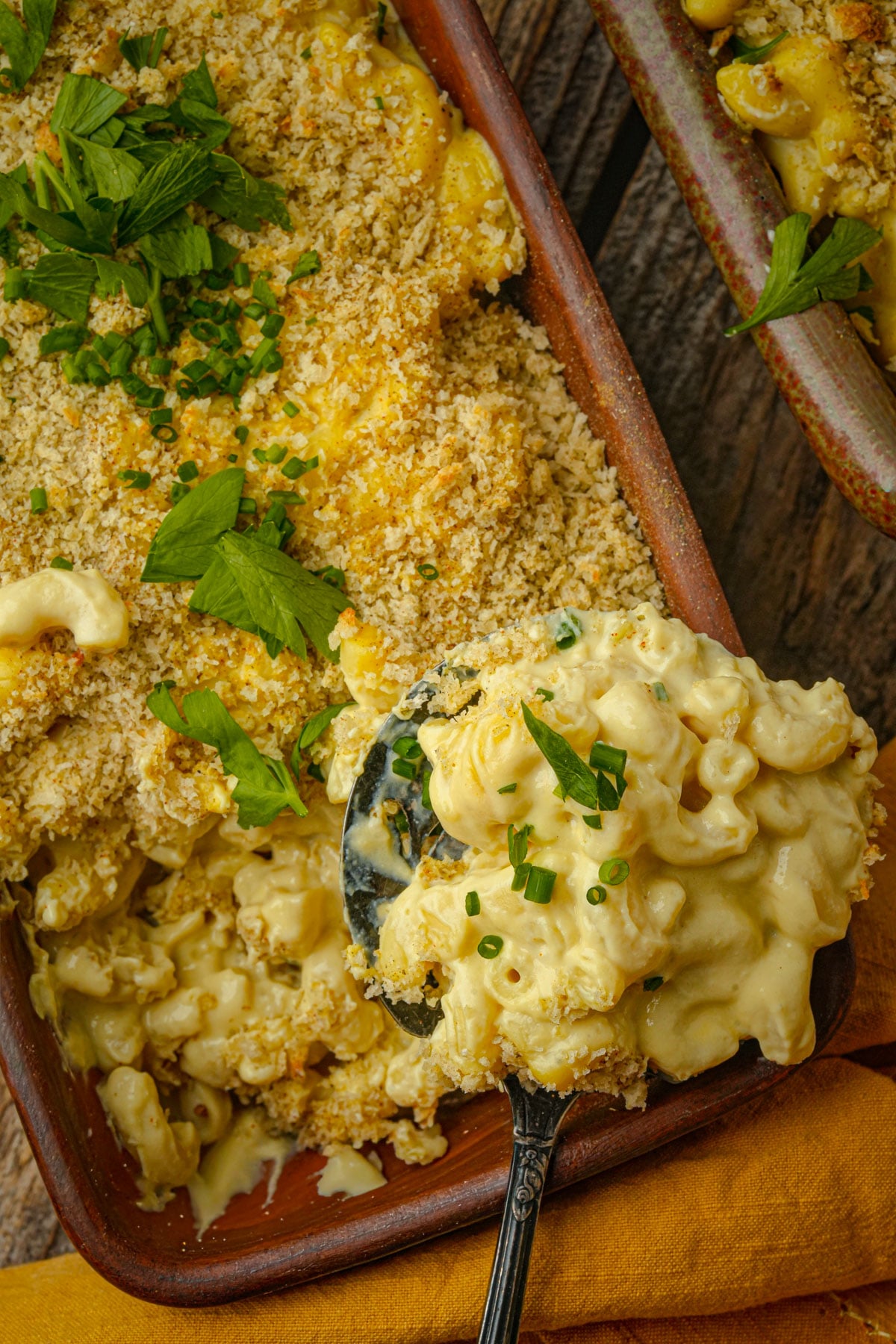 A serving spoon scoops creamy baked vegan truffle macaroni and cheese topped with golden breadcrumbs and garnished with fresh herbs from a rectangular dish.