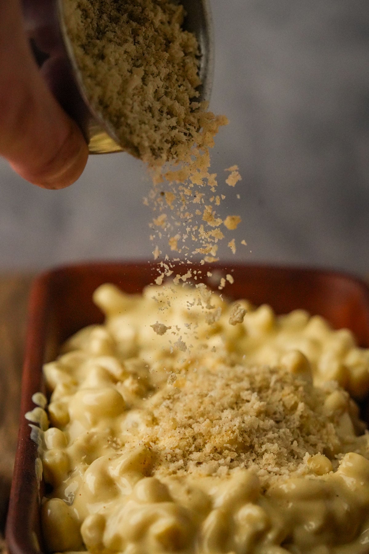 A close-up of a hand sprinkling breadcrumbs over a dish of creamy vegan truffle macaroni and cheese in a brown rectangular dish.