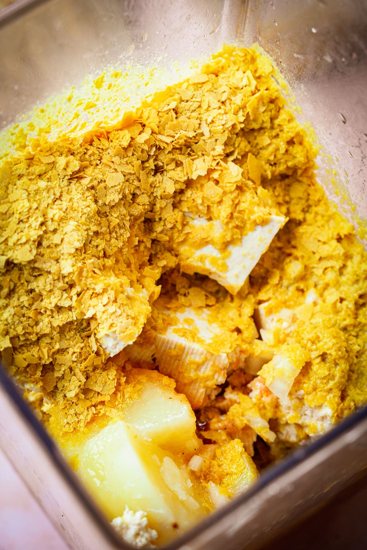 Close-up of a blender containing yellow nutritional yeast flakes, white tofu cubes, and potato pieces, ready to be blended for a recipe.