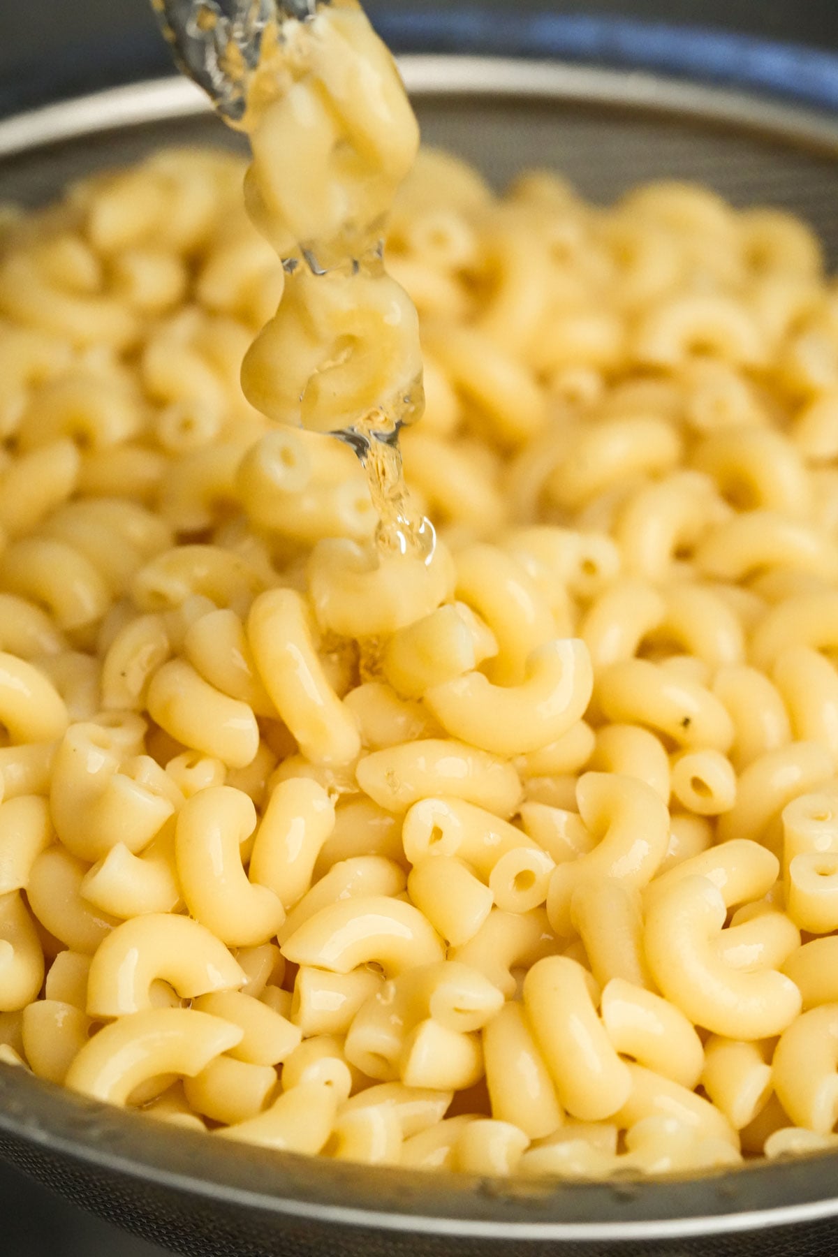 A close-up of cooked elbow macaroni being rinsed with water in a metal colander, with water droplets visible on the pasta.