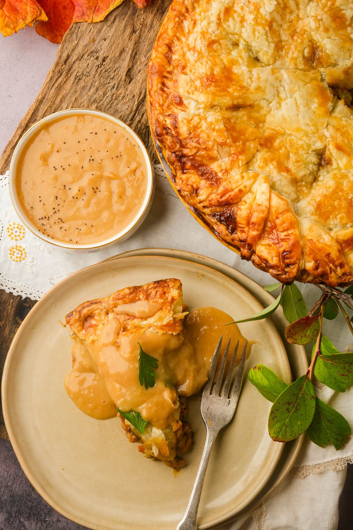 A slice of savory vegan pot pie with golden crust sits on a plate, covered in brown gravy, with a fork beside it. Nearby are a bowl of gravy and the rest of the pie, garnished with a green leaf.