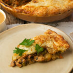 A slice of vegan pot pie with golden, flaky crust, filled with vegetables and soy curls, sits on a plate. The slice is garnished with fresh parsley, and the rest of the pie is visible in the background.