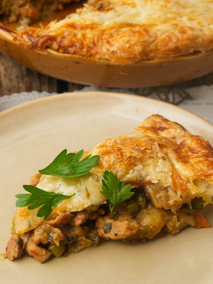 A slice of vegan pot pie with golden, flaky crust, filled with vegetables and soy curls, sits on a plate. The slice is garnished with fresh parsley, and the rest of the pie is visible in the background.