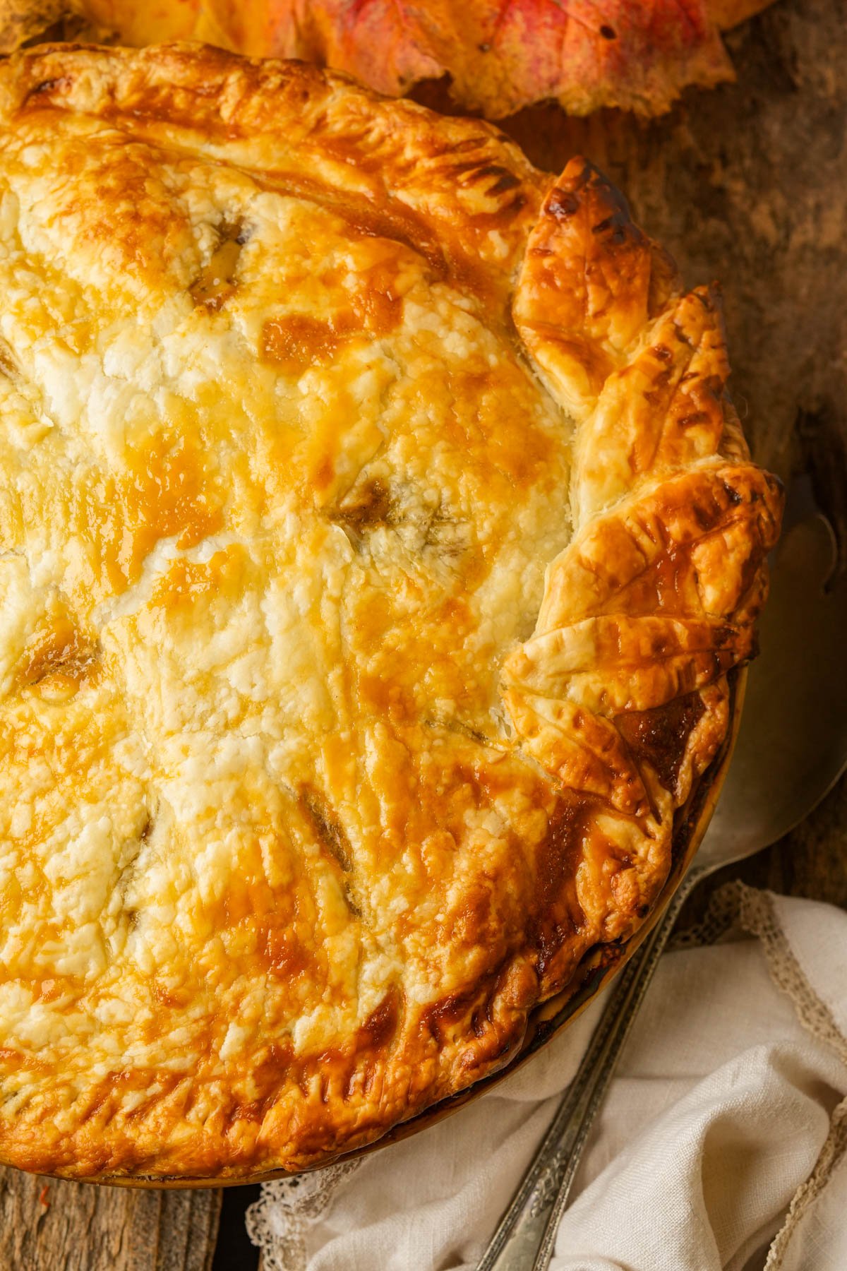 A golden-brown, homemade vegan pot pie with a flaky crust, sitting on a rustic wooden surface next to a white cloth and autumn leaves.