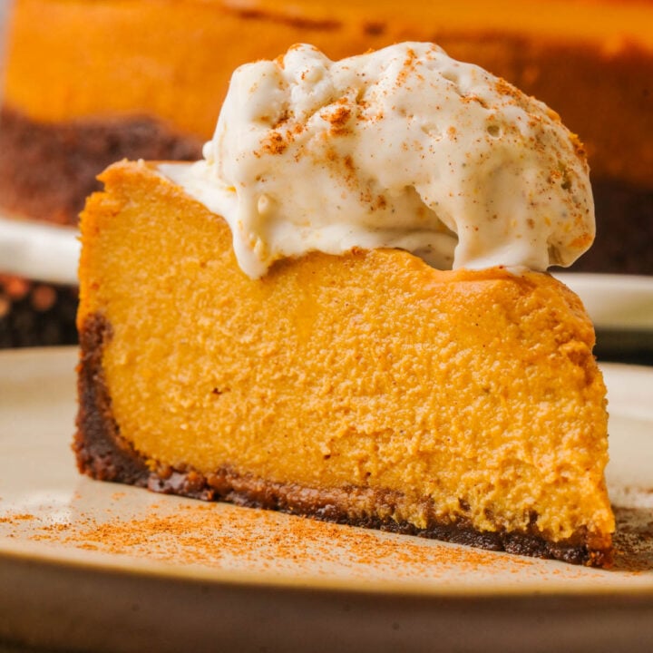 A slice of vegan pumpkin cheesecake on a plate, topped with a scoop of vanilla ice cream and sprinkled with cinnamon. The cheesecake has a brown crust and a creamy orange filling.