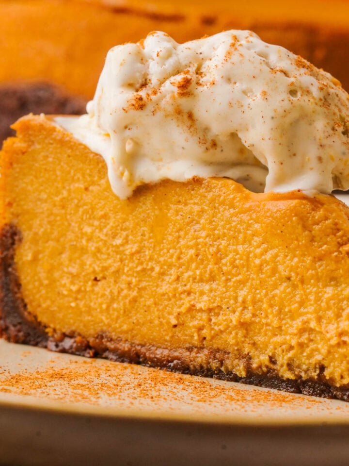 A slice of vegan pumpkin cheesecake on a plate, topped with a scoop of vanilla ice cream and sprinkled with cinnamon. The cheesecake has a brown crust and a creamy orange filling.