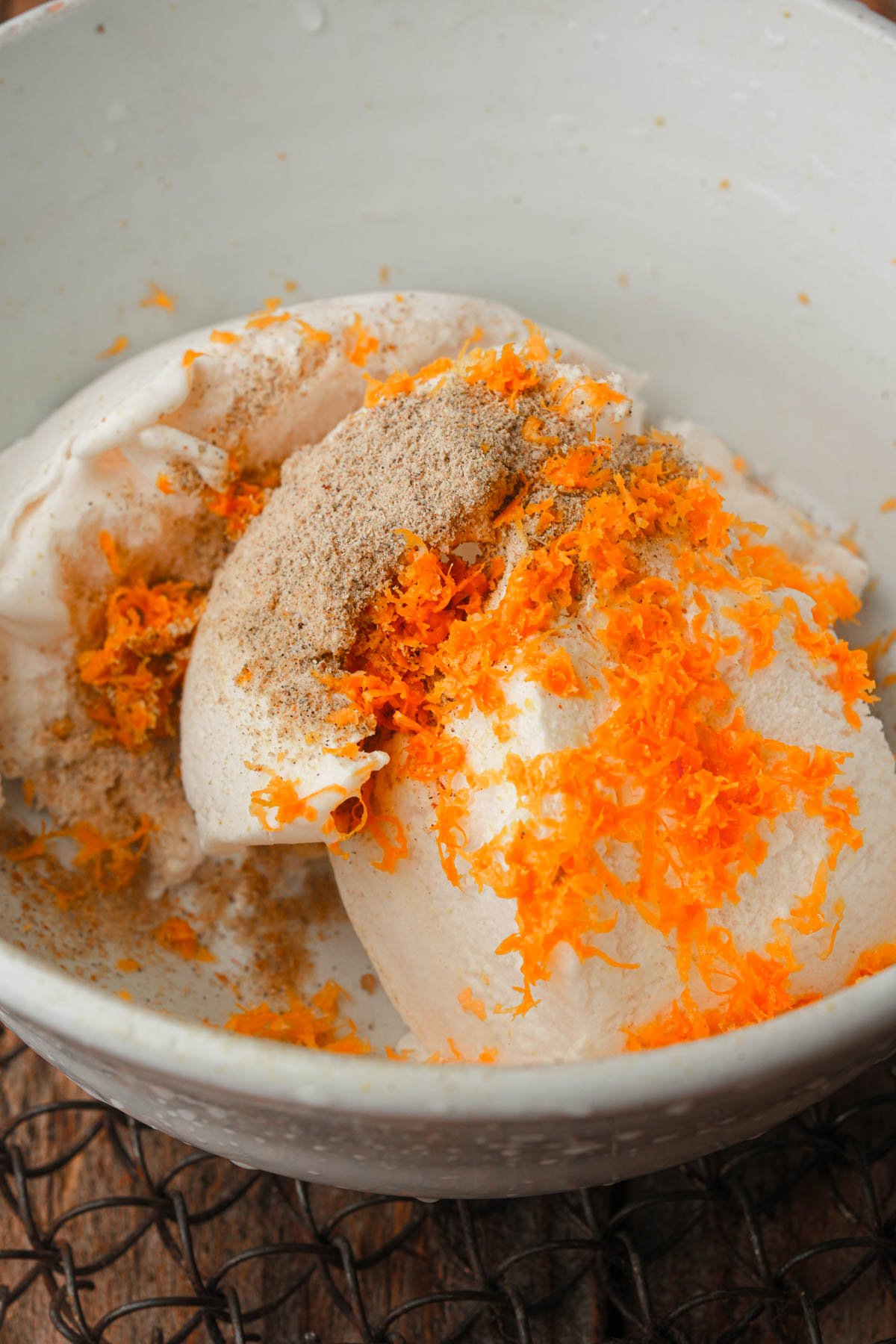 A white bowl filled with vegan whipped cream, topped with grated orange zest and sprinkled with ground cardamom, sitting on a wooden surface.