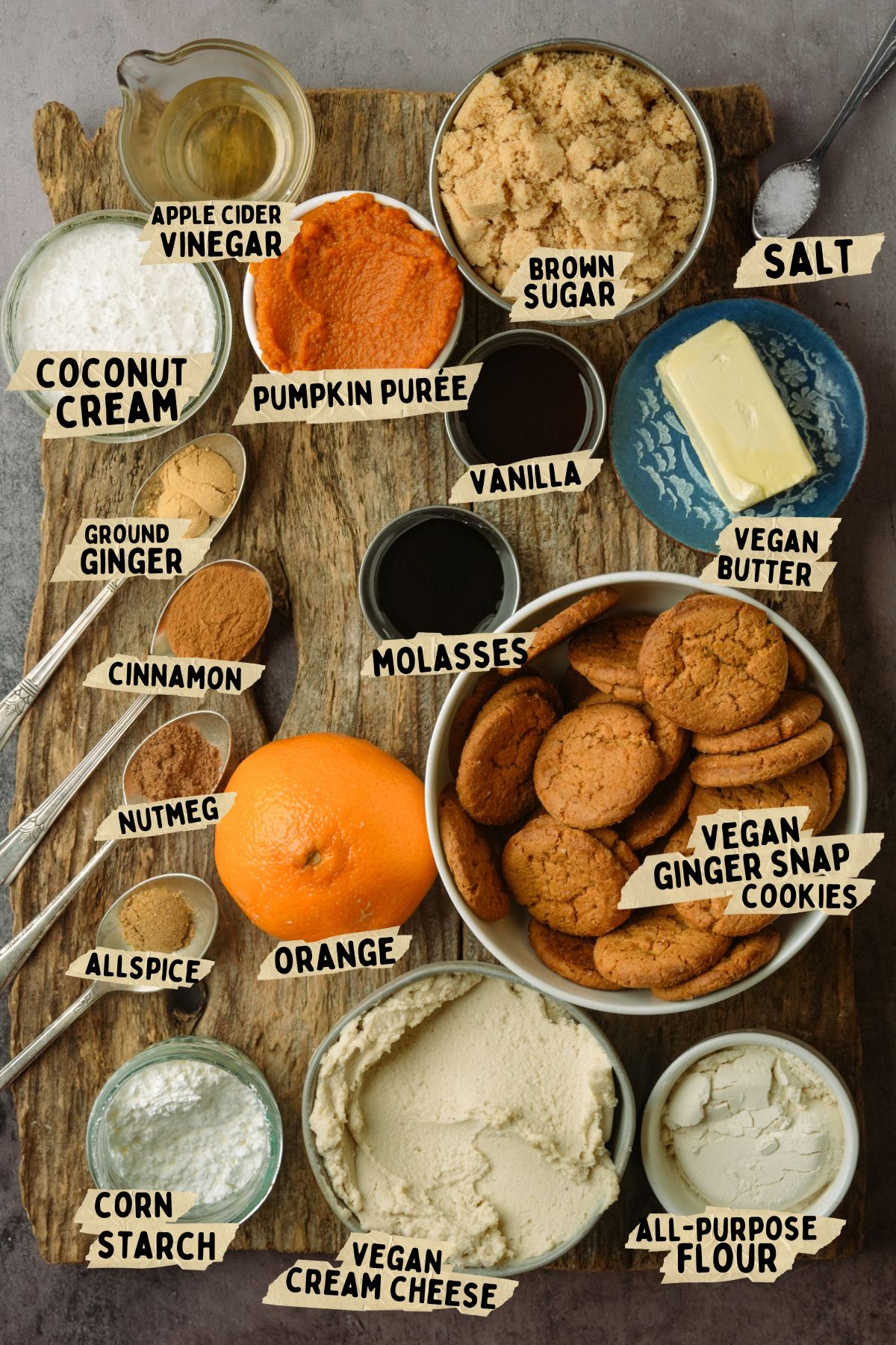 A rustic wooden board displays labeled baking ingredients for Vegan Pumpkin Cheesecake, including brown sugar, vegan butter, gingersnap cookies, pumpkin purée, orange, spices, vegan cream cheese, coconut cream, vinegar, vanilla, molasses, cornstarch, and flour.