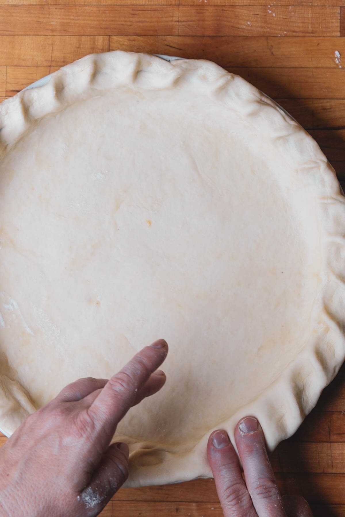 Two hands crimp the edges of an unbaked vegan pumpkin pie crust in a metal pie dish, resting on a wooden surface.
