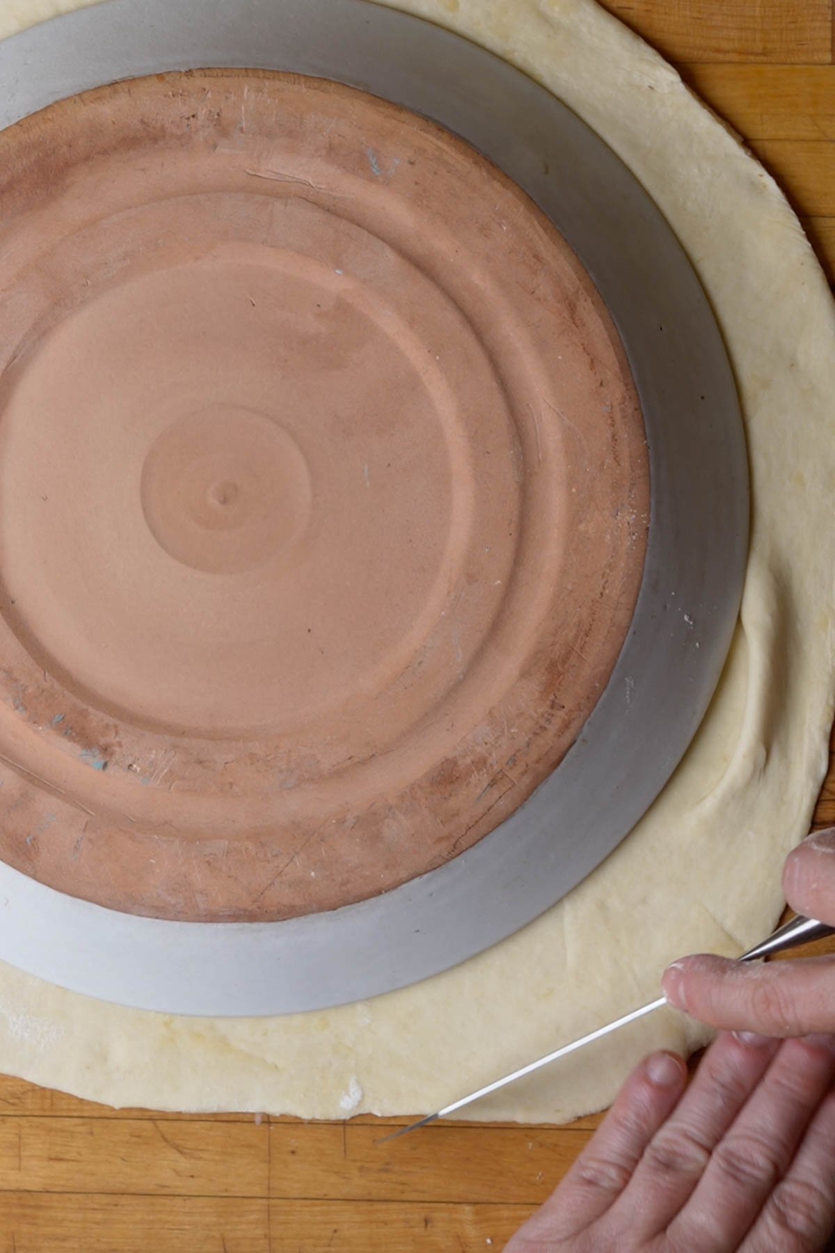A person uses a knife to cut dough around the edge of an upside-down baking stone and pan placed on top of the dough on a wooden surface.