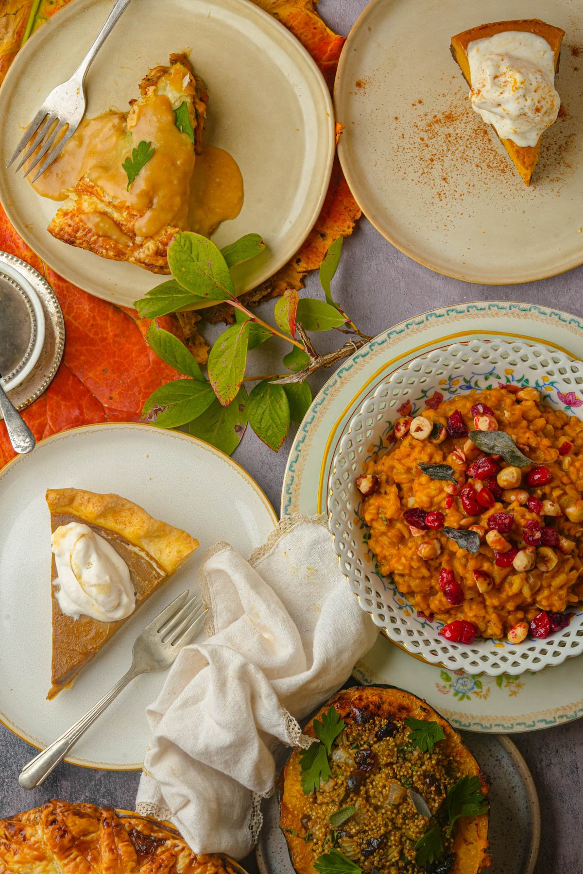 A table set with slices of vegan pumpkin pie topped with whipped cream, a bowl of pumpkin risotto with cranberries and hazelnuts, vegan stuffed acorn squash, vegan pumpkin cheesecake and a slice of vegan pot pit and autumn leaves for decoration.