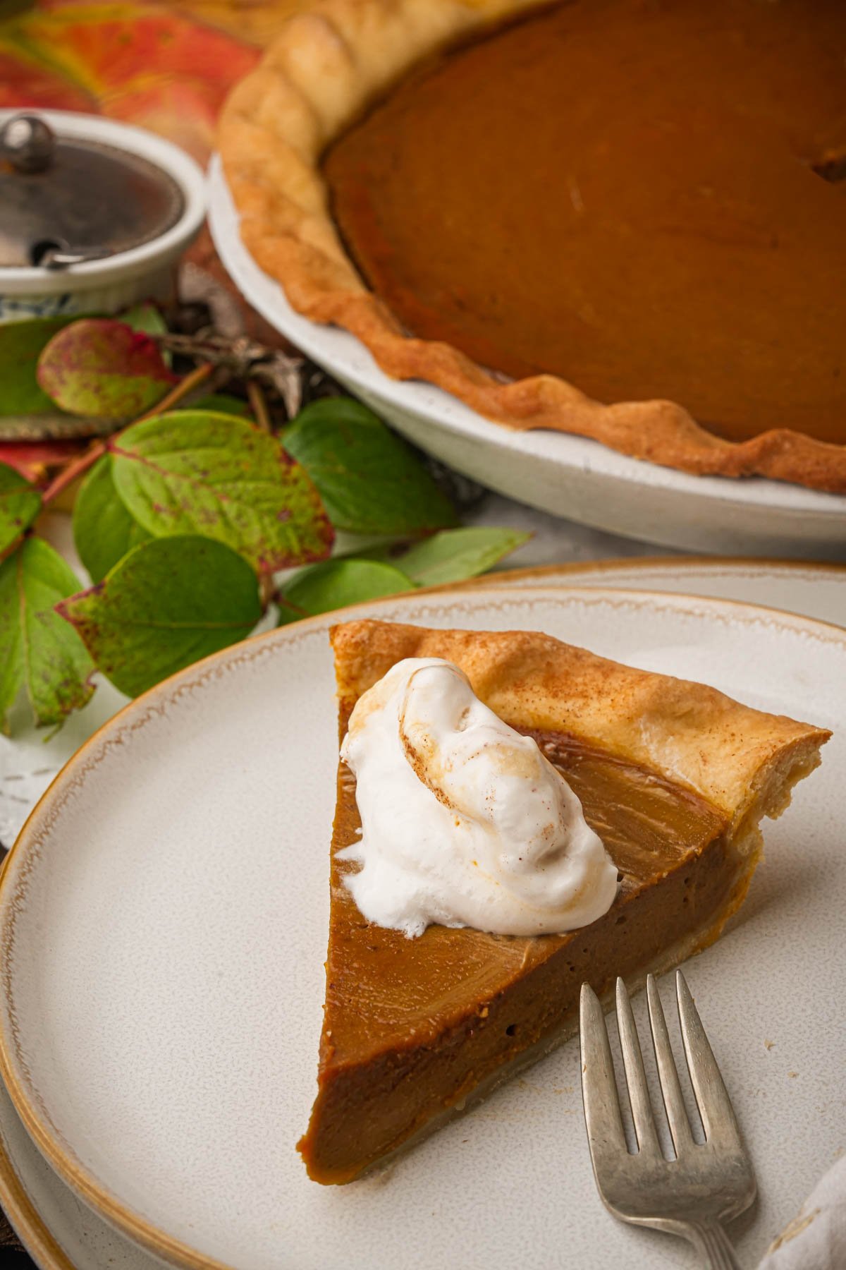 A slice of vegan pumpkin pie with a dollop of whipped cream sits on a plate with a fork, in front of a whole pumpkin pie. Autumn leaves and a sugar bowl are in the background.