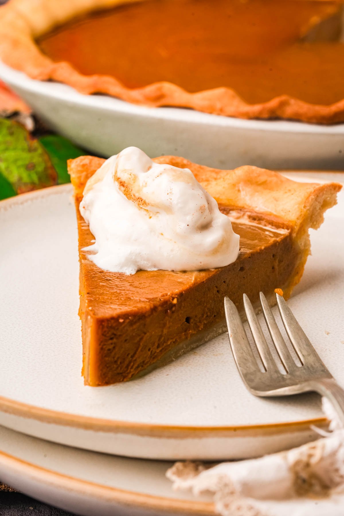 A slice of vegan pumpkin pie topped with a dollop of whipped cream sits on a plate with a fork, with the rest of the pie in the background.