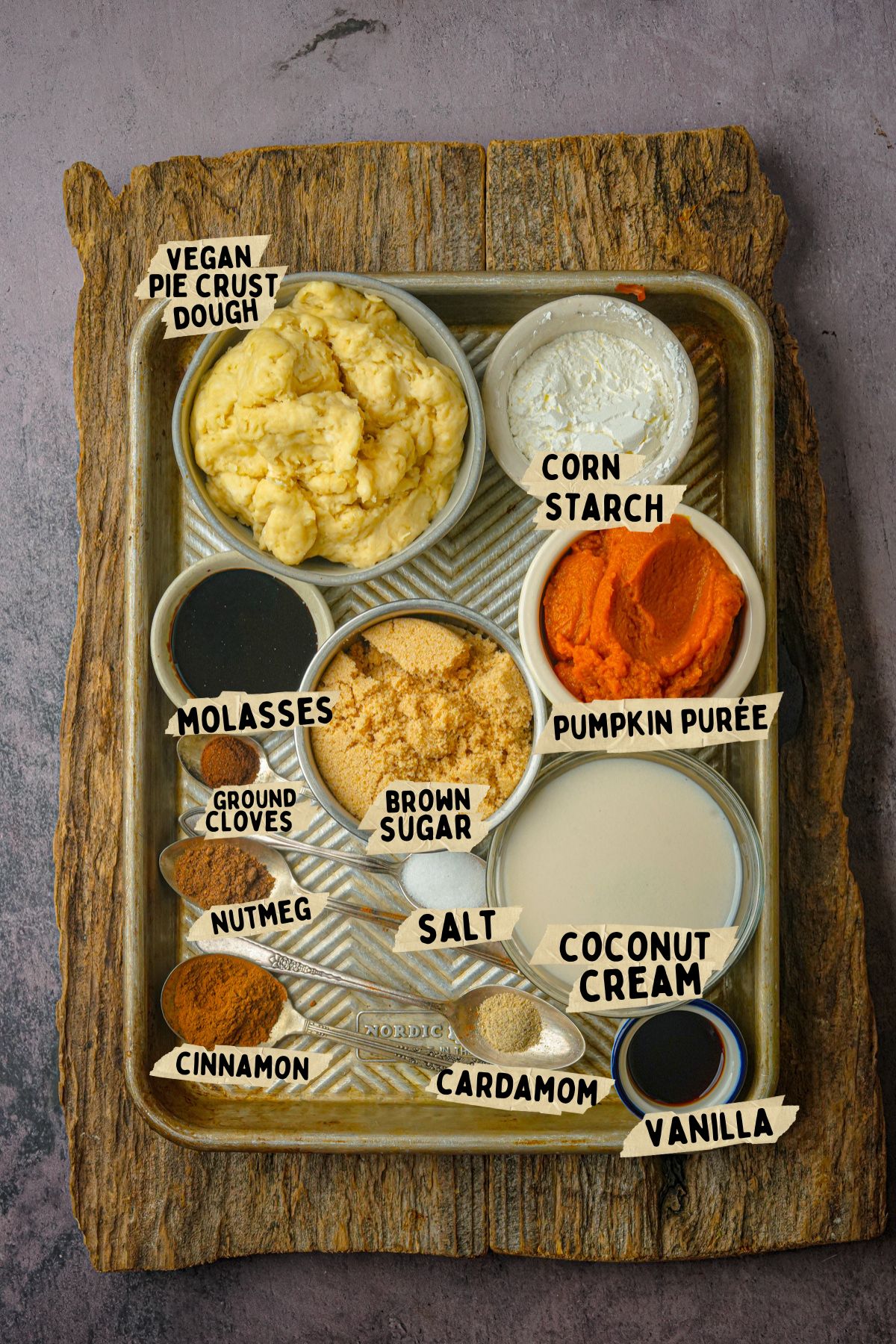 A tray on a wooden board holds bowls of ingredients for a vegan pumpkin pie recipe including vegan pie crust dough, cornstarch, pumpkin purée, coconut cream, brown sugar, molasses, salt, vanilla, cardamom, cinnamon, nutmeg, and ground cloves. Each ingredient is labeled.
