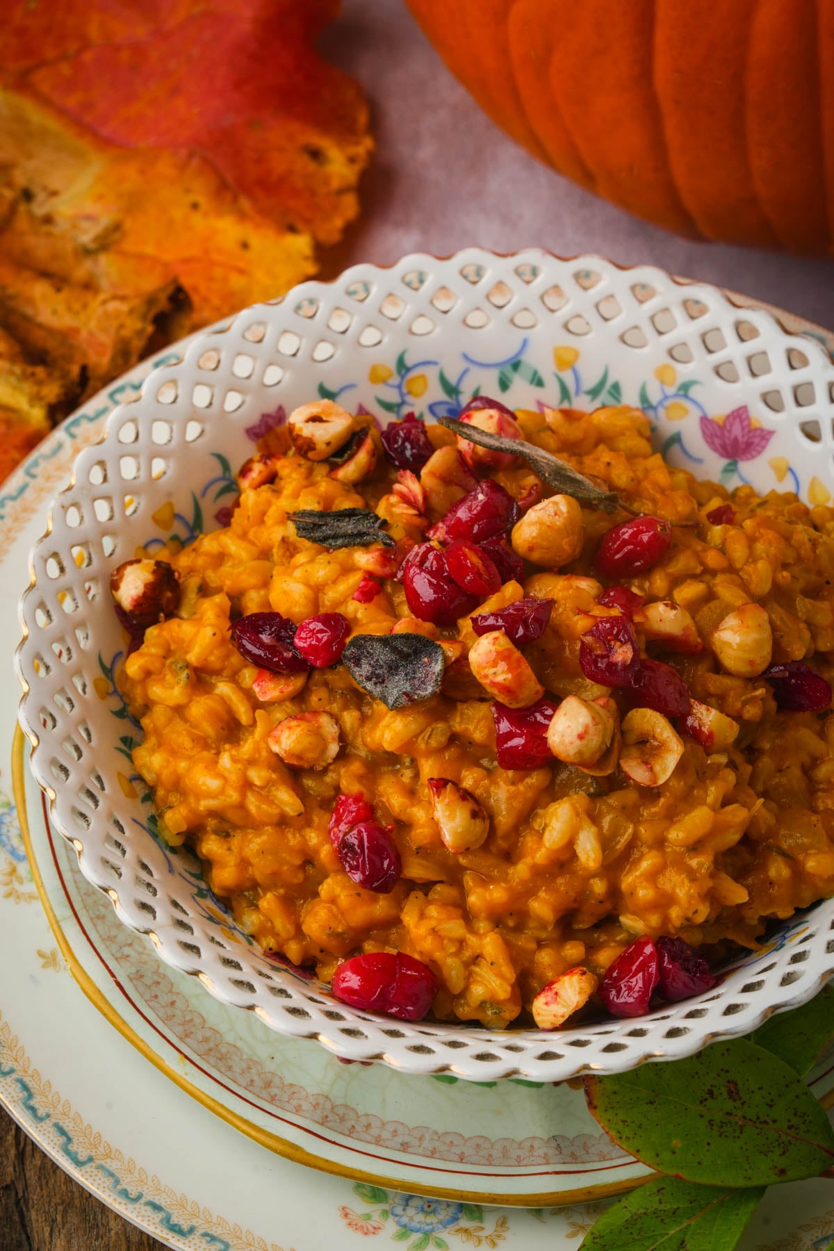 A bowl of pumpkin risotto topped with cranberries and hazelnuts sits on a decorative plate, surrounded by autumn leaves and a pumpkin, creating a cozy fall atmosphere.