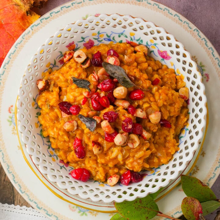 A bowl of creamy pumpkin risotto topped with toasted hazelnuts, dried cranberries, and sage leaves, served on a decorative floral plate with autumn leaves nearby.