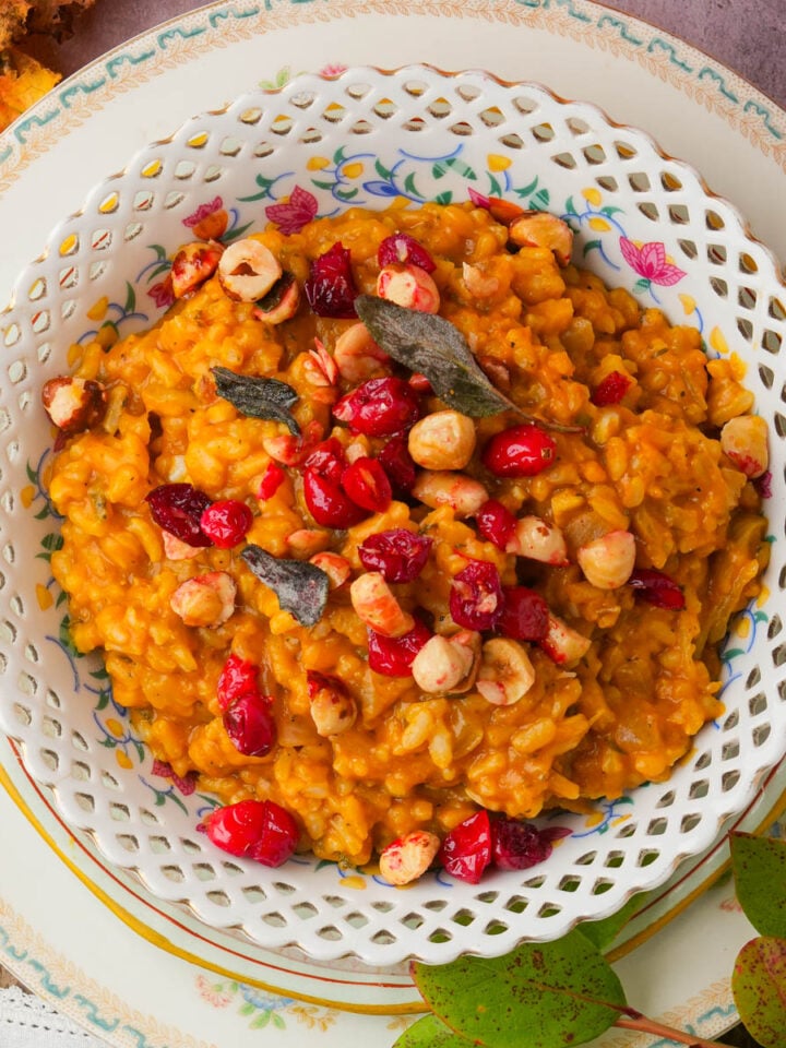A bowl of creamy pumpkin risotto topped with toasted hazelnuts, dried cranberries, and sage leaves, served on a decorative floral plate with autumn leaves nearby.