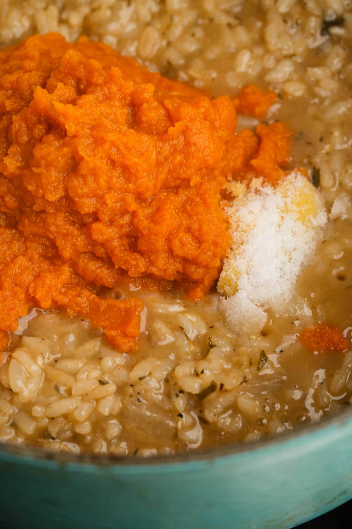 A close-up of creamy pumpkin risotto in a blue pan, topped with a generous scoop of orange pumpkin puree and a small spoonful of salt.