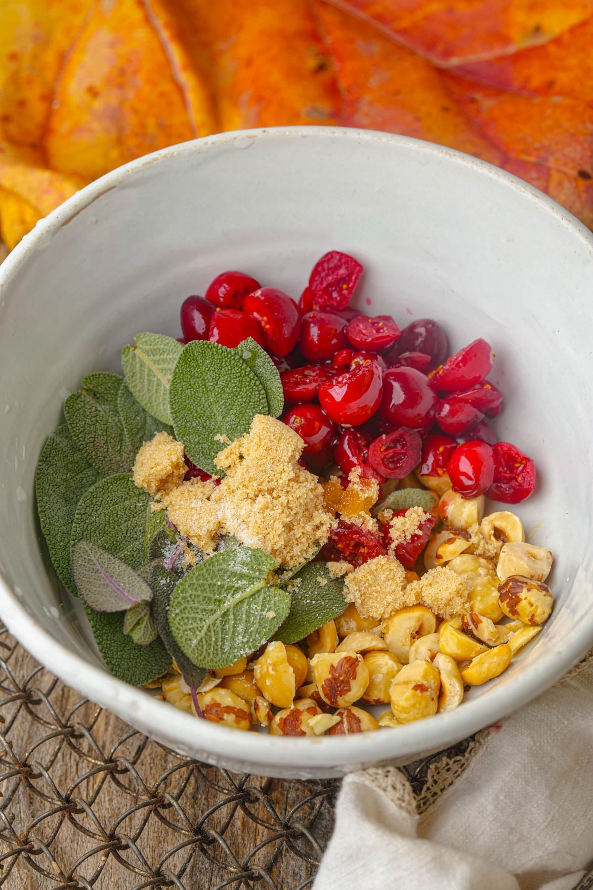 A white bowl containing fresh sage leaves, chopped cranberries, roasted hazelnuts, and a mound of brown sugar, set against a background with autumn leaves.