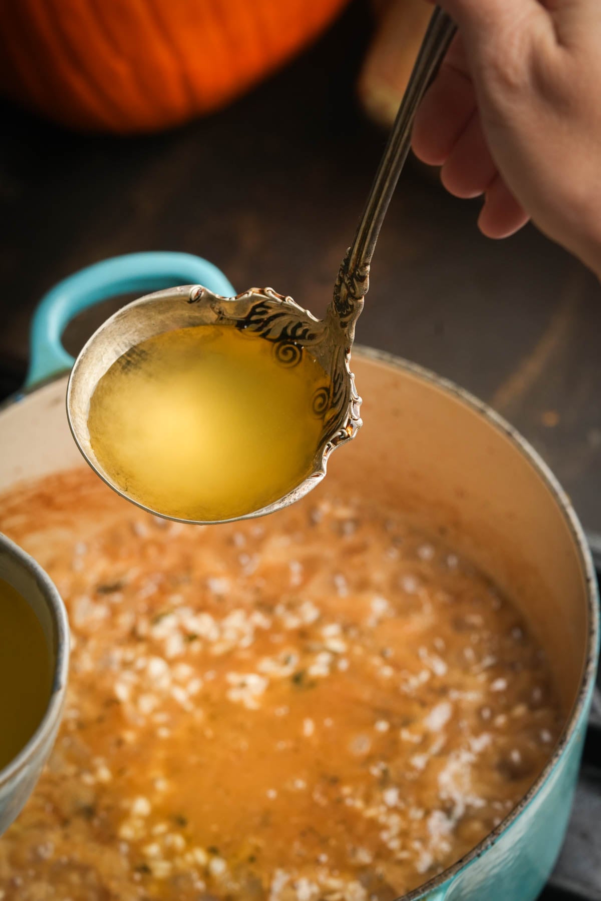 A hand holds an ornate ladle filled with clear stock above a pot of pumpkin risotto.