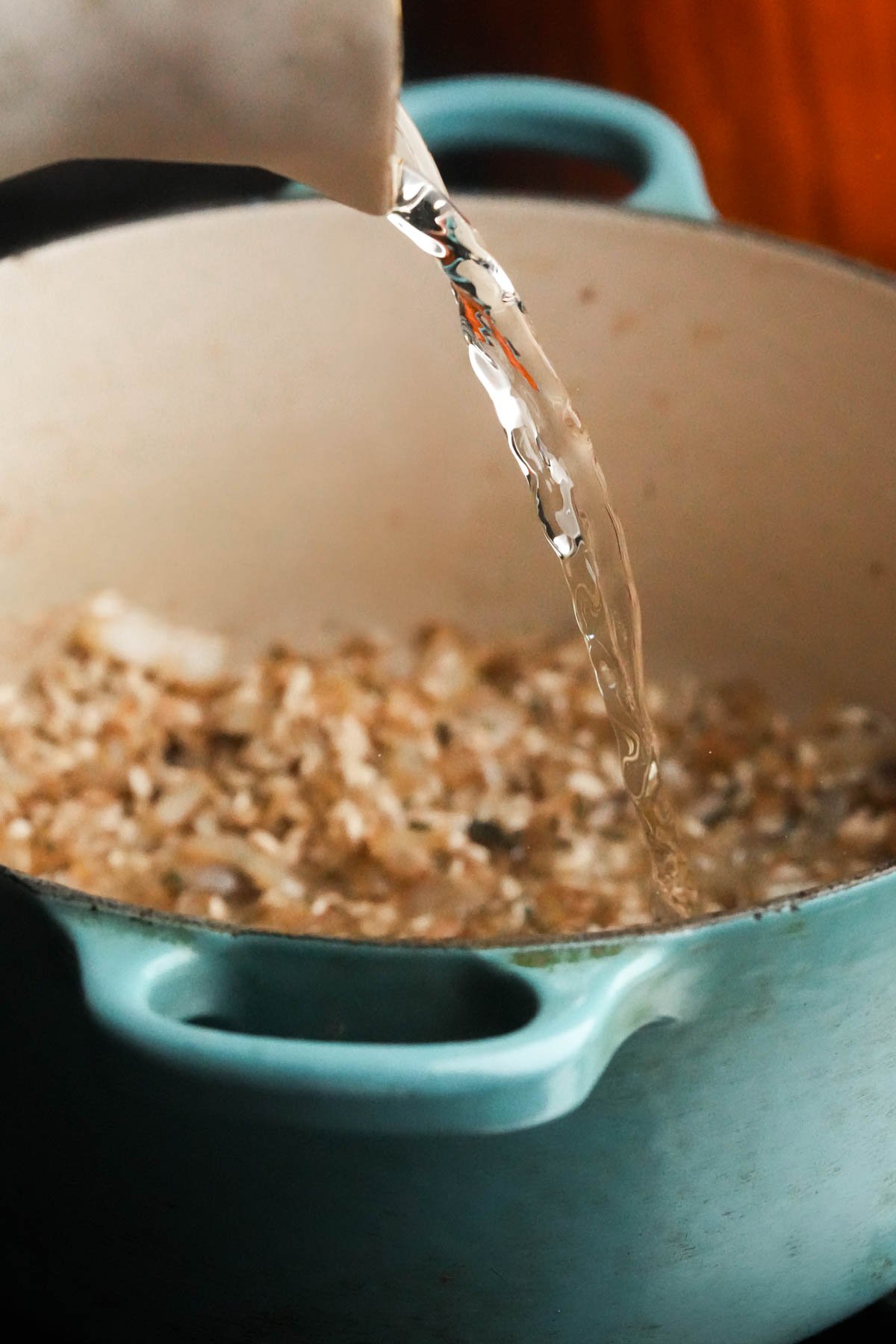White wine is being poured from a white pitcher into a light blue pot containing rice, chopped onions, and garlic.