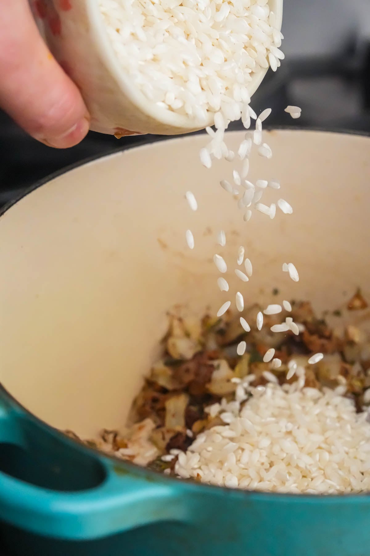 A hand pours uncooked arborio rice from a container into a teal pot with sautéed vegetables inside.