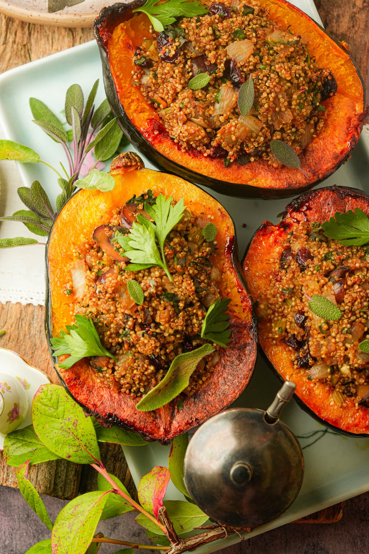 Three vegan stuffed acorn squash halves stuffed with a quinoa and vegetable mixture, garnished with fresh herbs, are arranged on a tray with a metal oil dispenser and scattered green leaves.